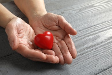 Photo of Elderly woman holding red heart in hands at grey wooden table, closeup
