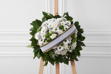 Photo of Funeral wreath of flowers with ribbon on wooden stand near white wall indoors
