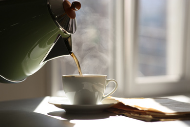 Photo of Pouring hot drink into cup near newspaper on sunlit table. Good morning