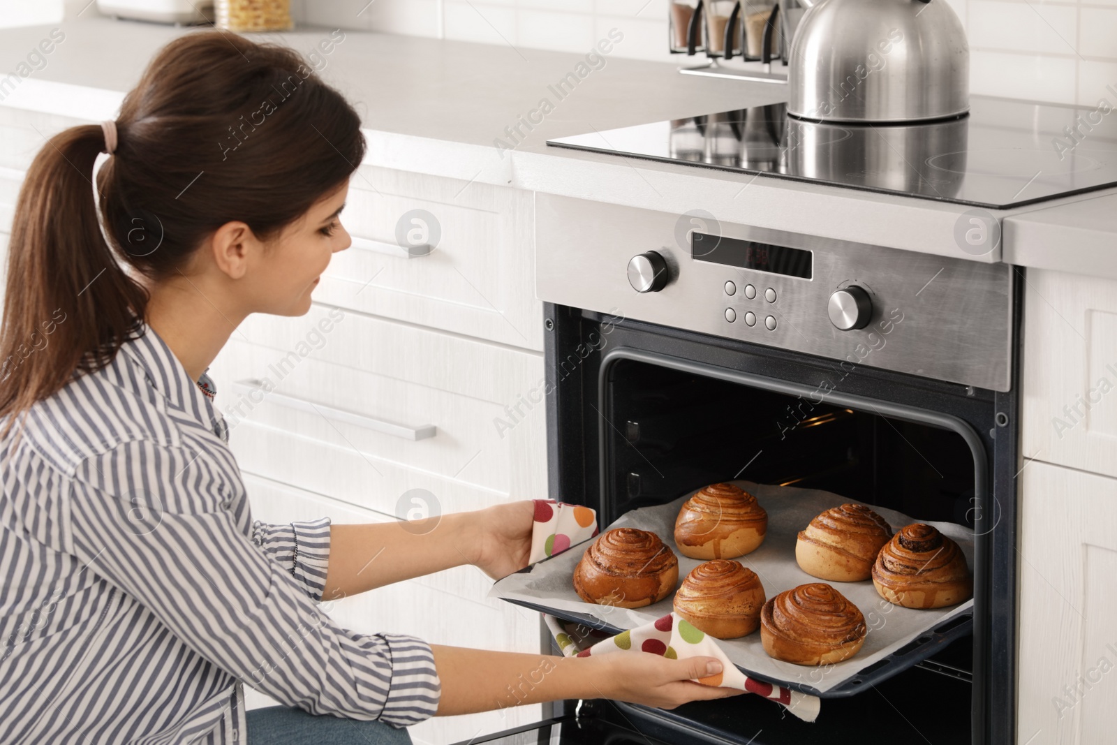 Photo of Beautiful young woman taking out tray of baked buns from oven in kitchen