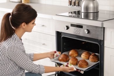 Photo of Beautiful young woman taking out tray of baked buns from oven in kitchen