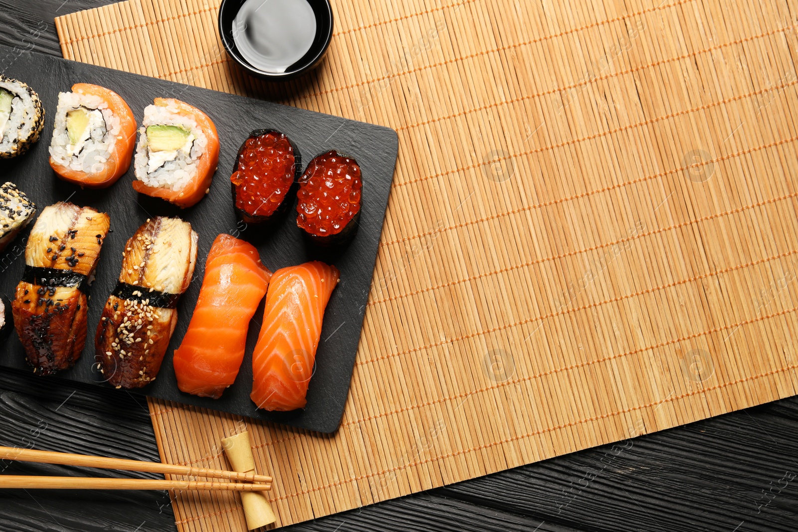 Photo of Delicious sushi rolls, soy sauce and chopsticks on black wooden table, flat lay. Space for text