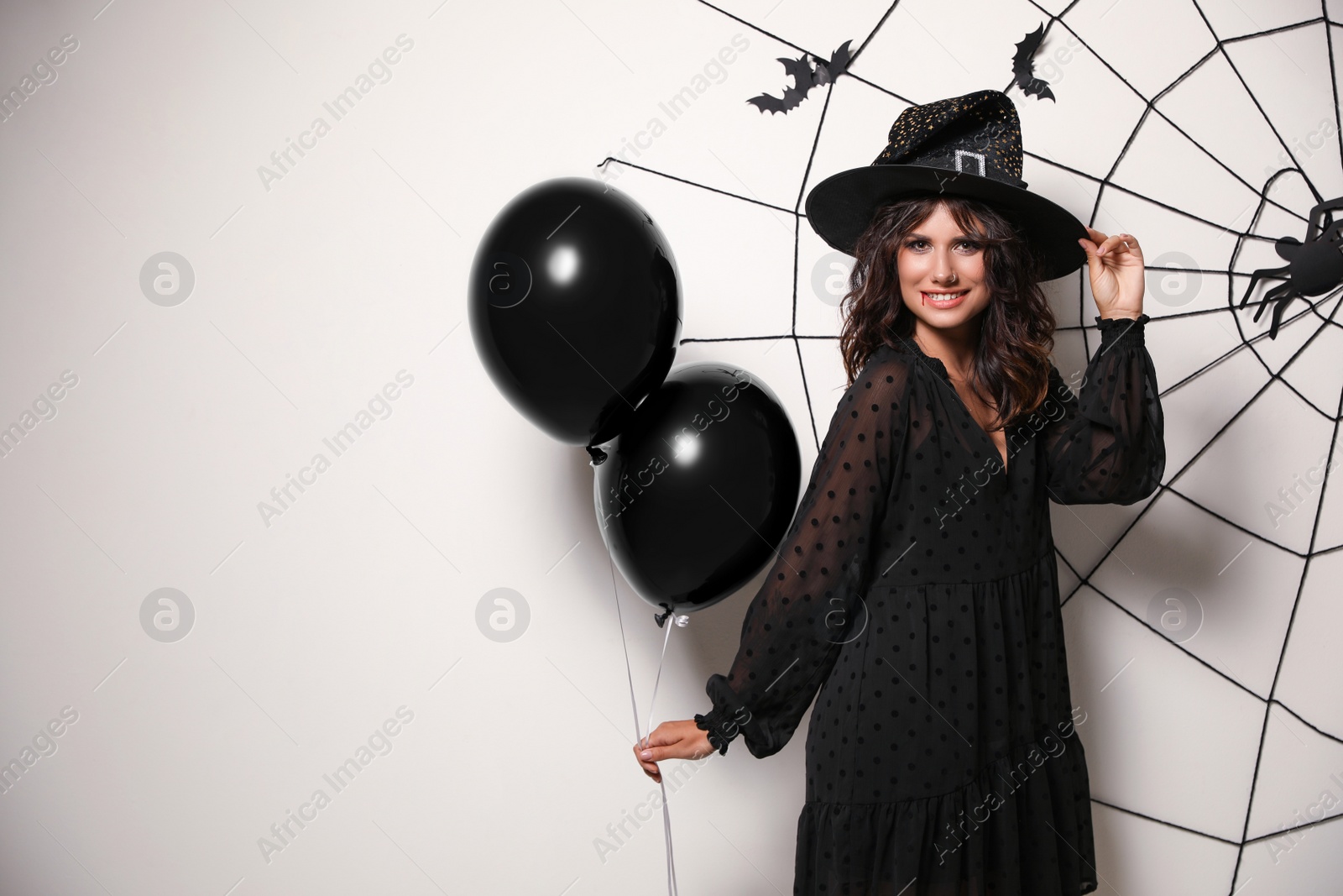 Photo of Woman in witch hat with balloons posing near white wall decorated for Halloween