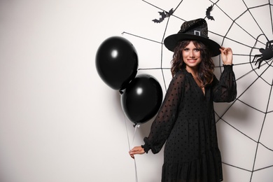 Photo of Woman in witch hat with balloons posing near white wall decorated for Halloween