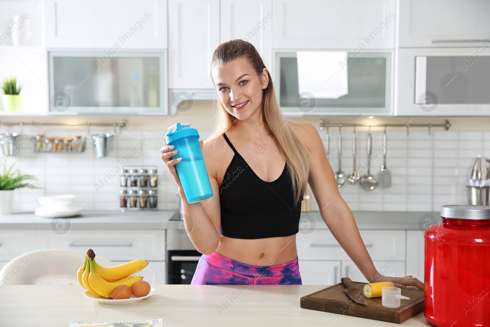 Photo of Young woman holding bottle of protein shake near table with ingredients in kitchen