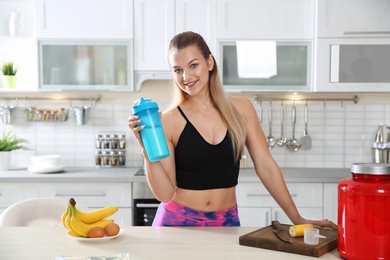 Photo of Young woman holding bottle of protein shake near table with ingredients in kitchen