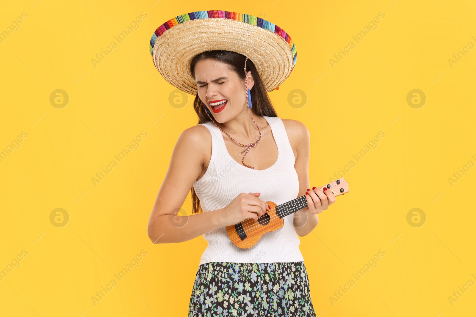 Photo of Young woman in Mexican sombrero hat playing ukulele on yellow background