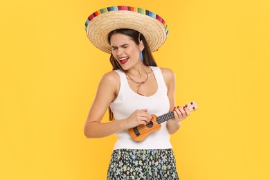 Photo of Young woman in Mexican sombrero hat playing ukulele on yellow background