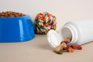 Bottle with vitamins, toy and dry pet food in bowl on beige table, closeup. Space for text