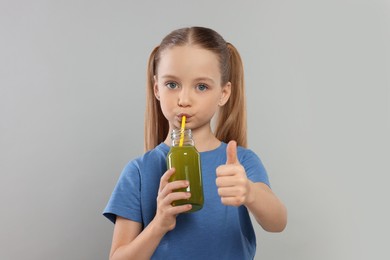 Cute little girl drinking fresh juice and showing thumbs up on light gray background
