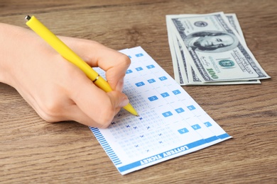 Photo of Woman filling out lottery tickets with pen and money on wooden table, closeup