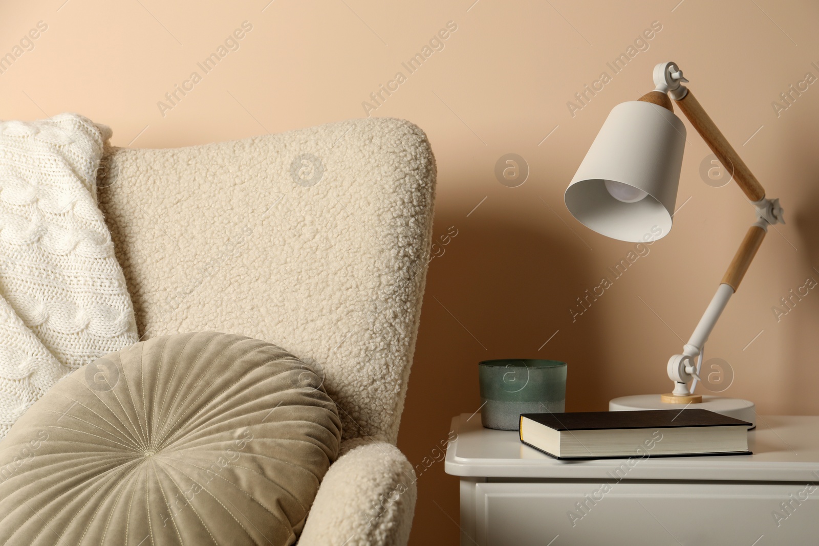 Photo of Stylish modern desk lamp, book and candle on white cabinet near soft armchair indoors