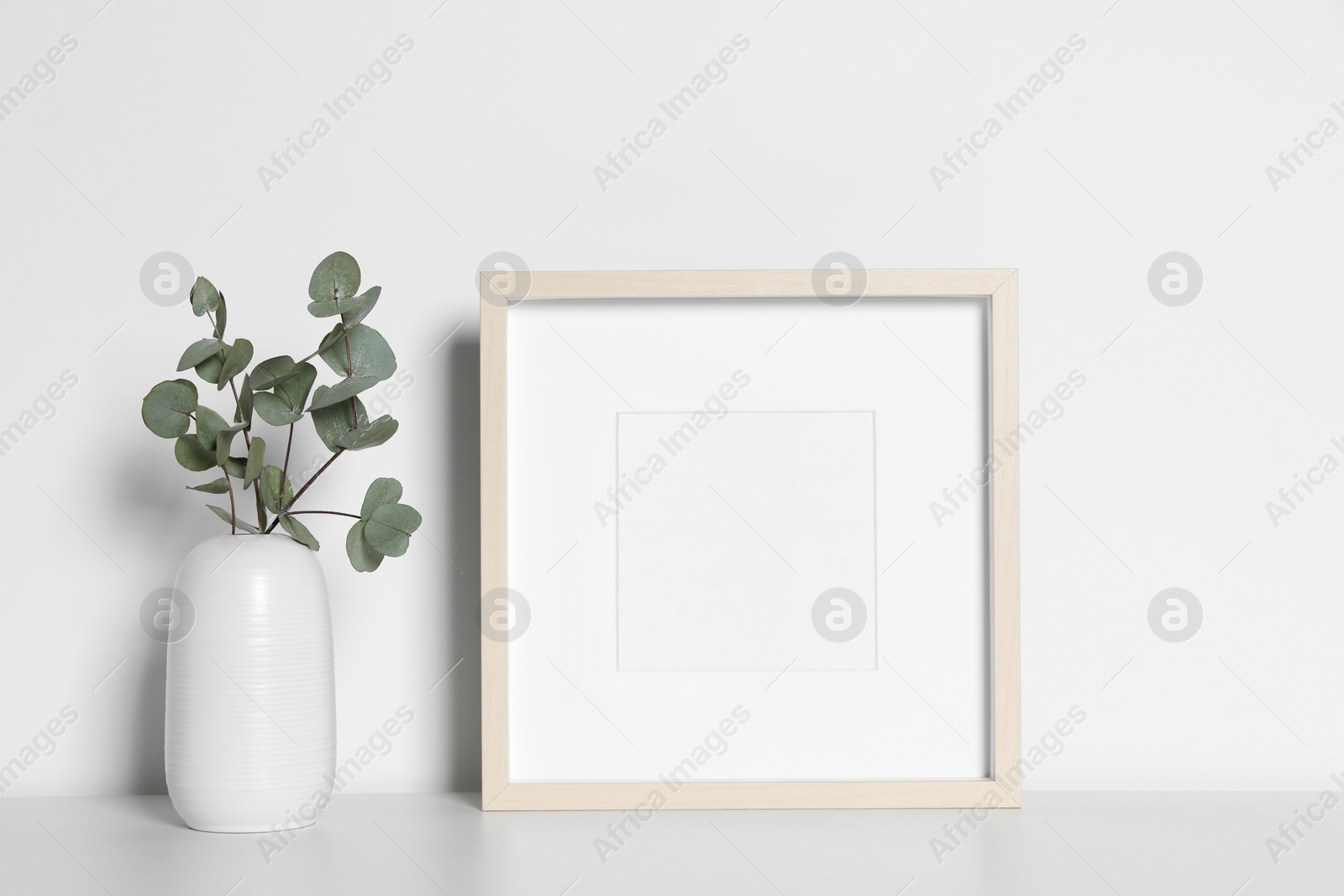 Photo of Empty photo frame and vase with decorative eucalyptus leaves on white table. Mockup for design