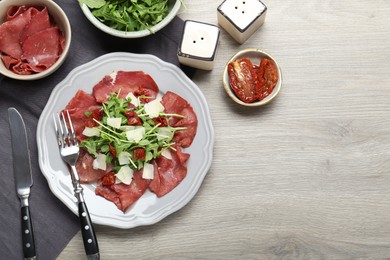 Photo of Plate of tasty bresaola salad with sun-dried tomatoes, parmesan cheese and cutlery on wooden table, flat lay. Space for text
