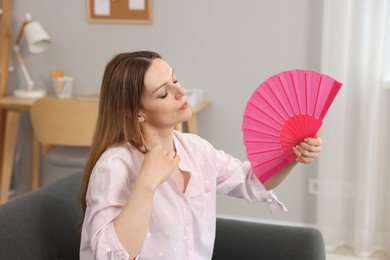 Photo of Woman waving pink hand fan to cool herself on sofa at home