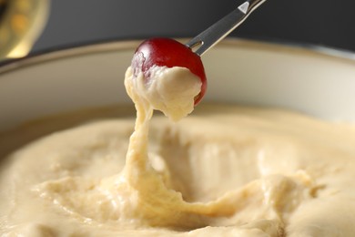 Photo of Dipping piece of grape into fondue pot with melted cheese on grey background, closeup