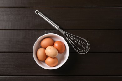 Photo of Metal whisk and raw eggs in bowl on dark wooden table, top view