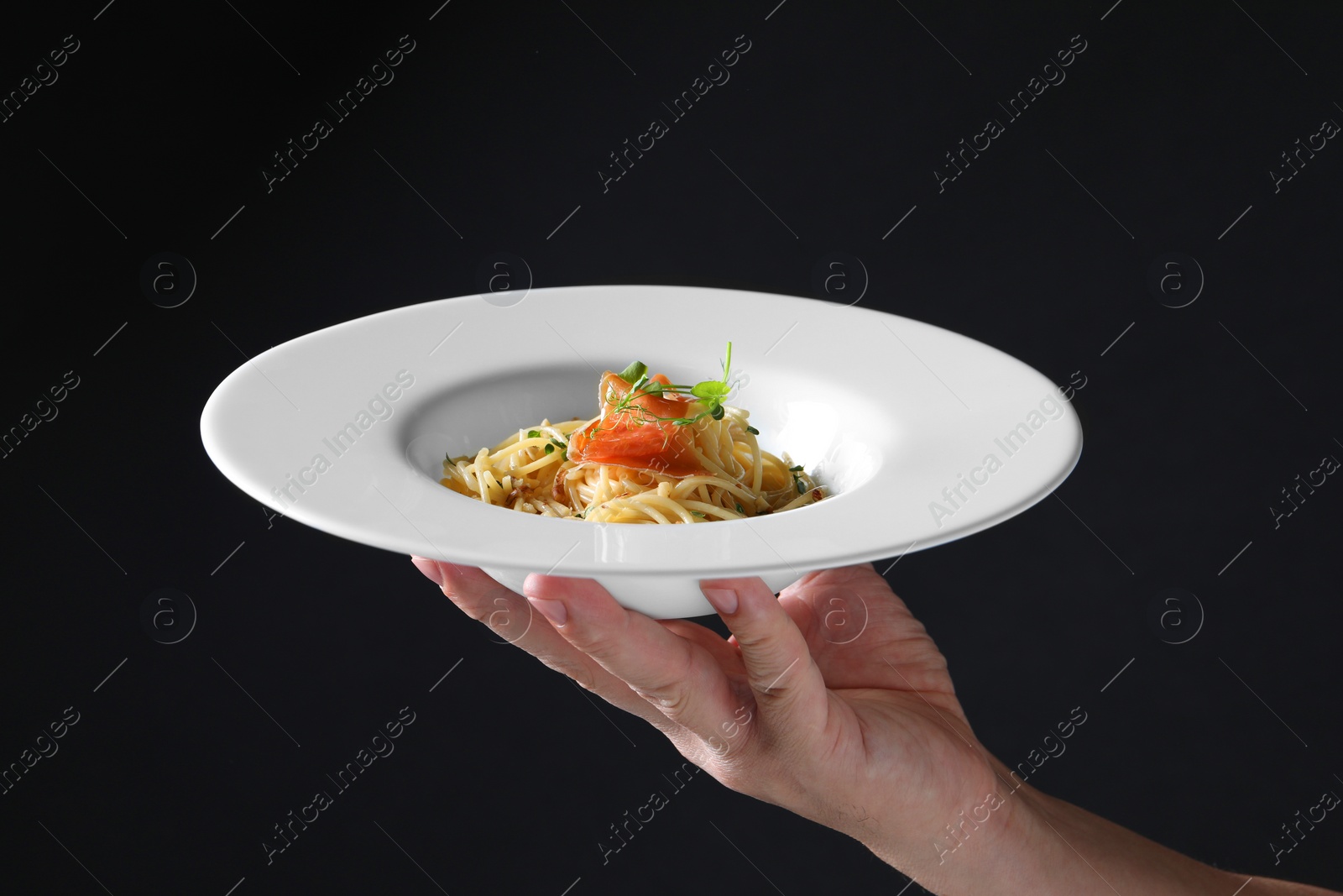 Photo of Woman holding plate of tasty spaghetti with prosciutto and microgreens on black background, closeup. Exquisite presentation of pasta dish