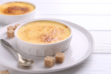 Delicious creme brulee in bowls, sugar cubes and spoon on white wooden table, closeup