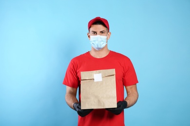 Photo of Courier in medical mask holding paper bag with takeaway food on light blue background. Delivery service during quarantine due to Covid-19 outbreak