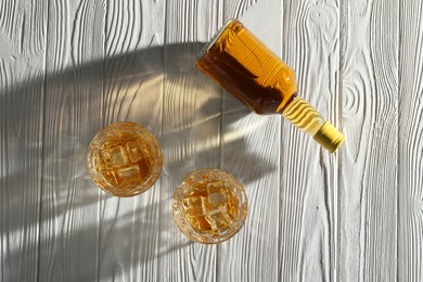 Photo of Whiskey with ice cubes in glasses and bottle on white wooden table, flat lay