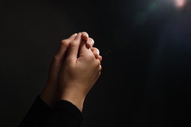 Photo of Woman holding hands clasped while praying against light in darkness, closeup. Space for text