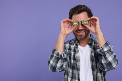 Photo of Man holding halves of kiwi near his eyes on purple background, space for text