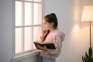 Photo of Cute little girl reading book near window at home