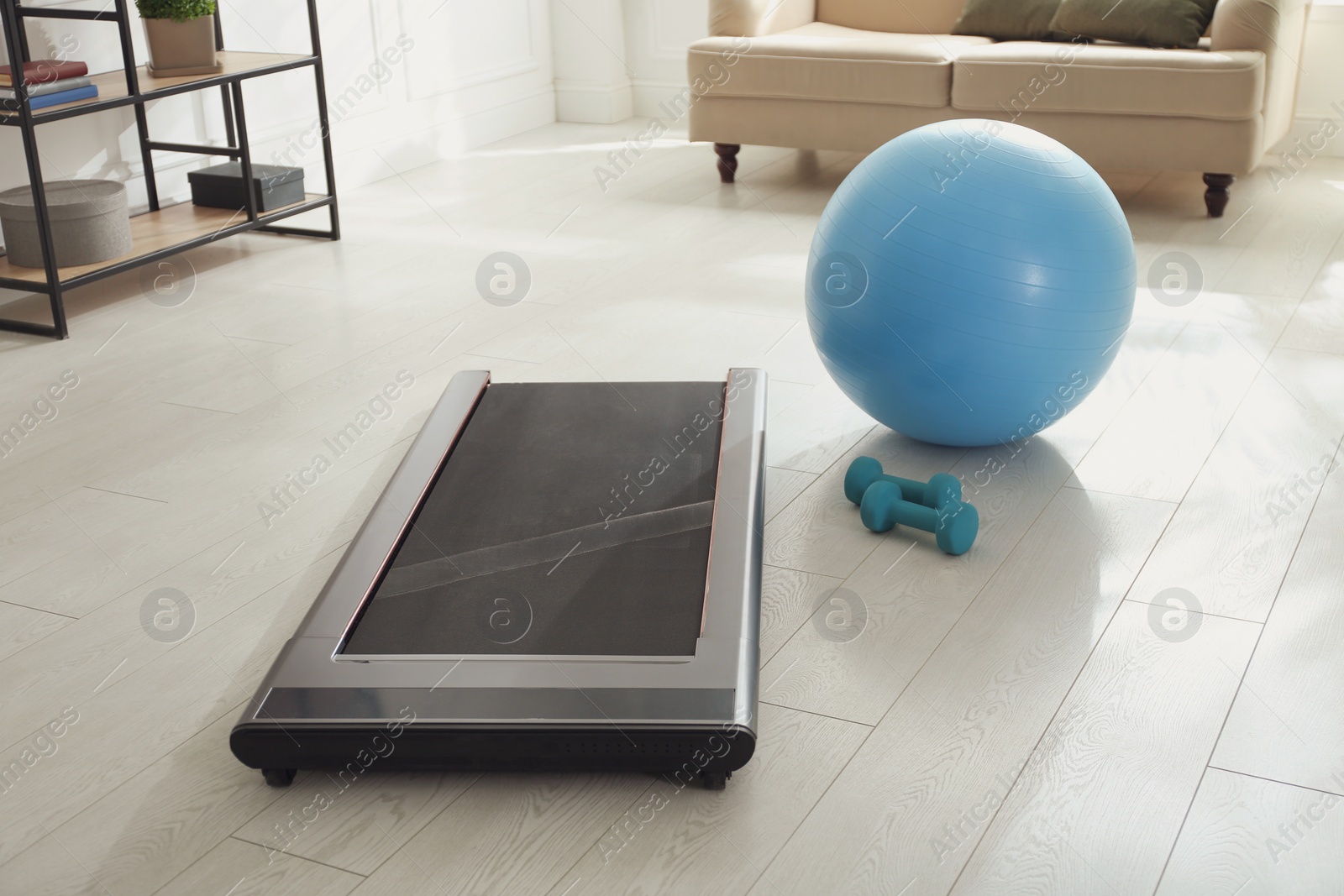 Photo of Modern walking treadmill, dumbbells and fitness ball in living room. Home gym equipment