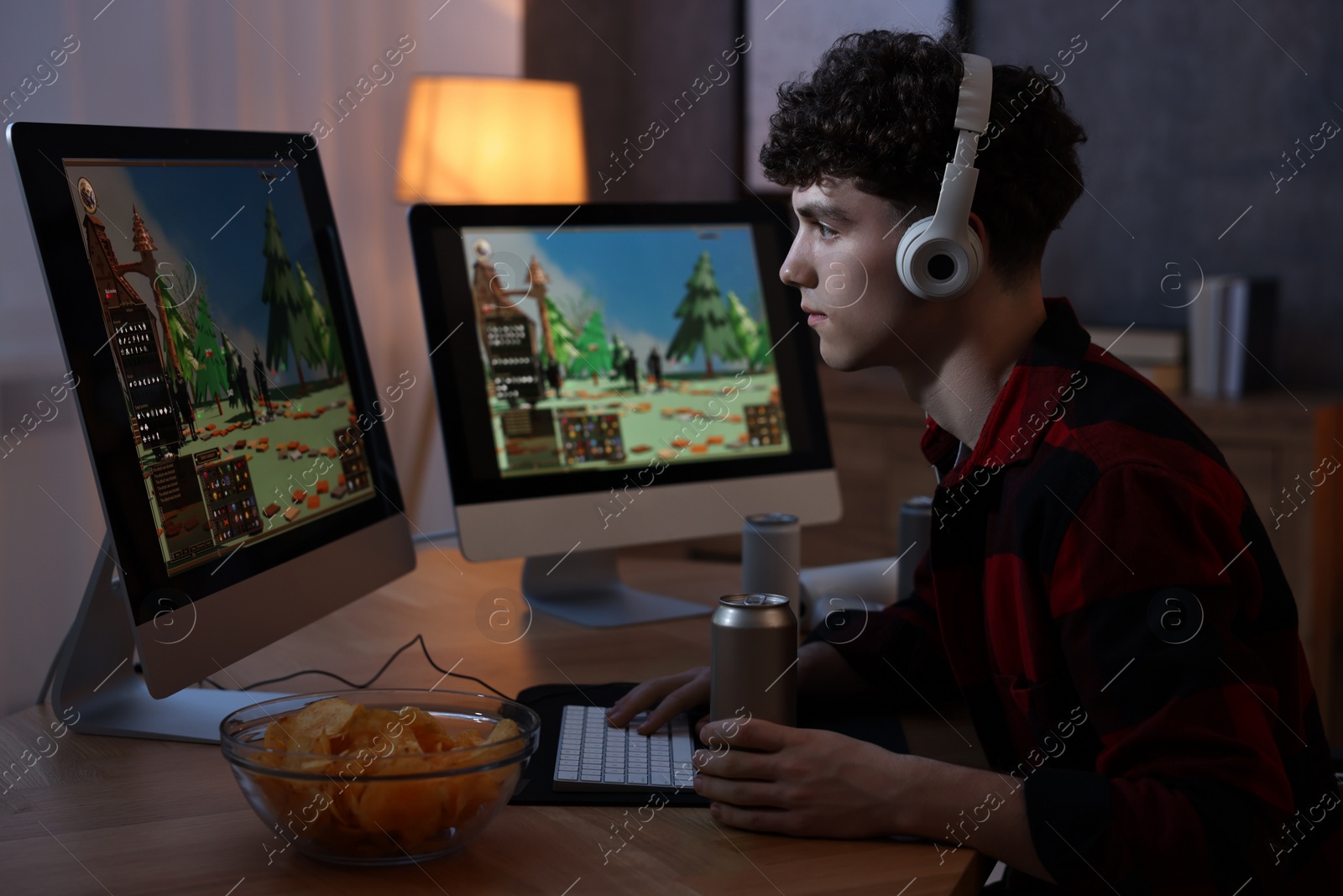 Photo of Young man with energy drink and headphones playing video game at wooden desk indoors