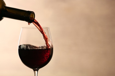 Pouring red wine into glass from bottle against blurred beige background, closeup. Space for text