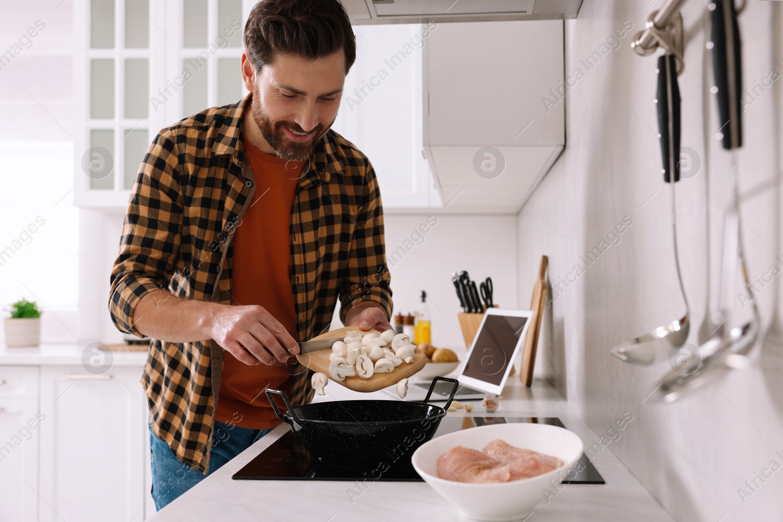 Photo of Man putting cut mushrooms into frying pan while watching online cooking course via laptop in kitchen