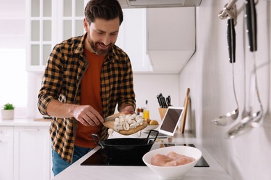 Man putting cut mushrooms into frying pan while watching online cooking course via laptop in kitchen