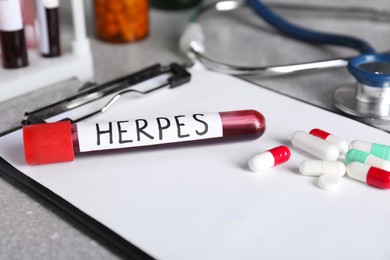 Test tube with word Herpes, different pills and clipboard on light grey table