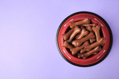 Photo of Red bowl with bone shaped dog cookies on purple background, top view. Space for text