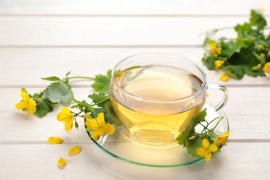 Photo of Glass cup of aromatic celandine tea and flowers on white wooden table