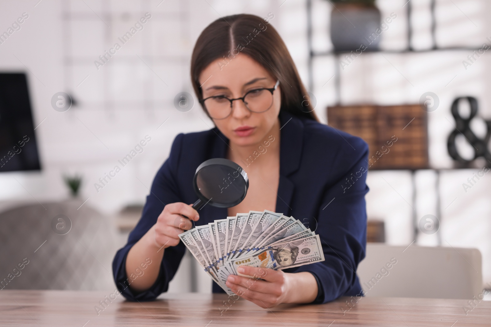 Photo of Expert authenticating 100 dollar banknotes with magnifying glass at table in office, focus on hand. Fake money concept