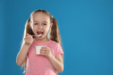 Cute little girl eating tasty yogurt on blue background. Space for text