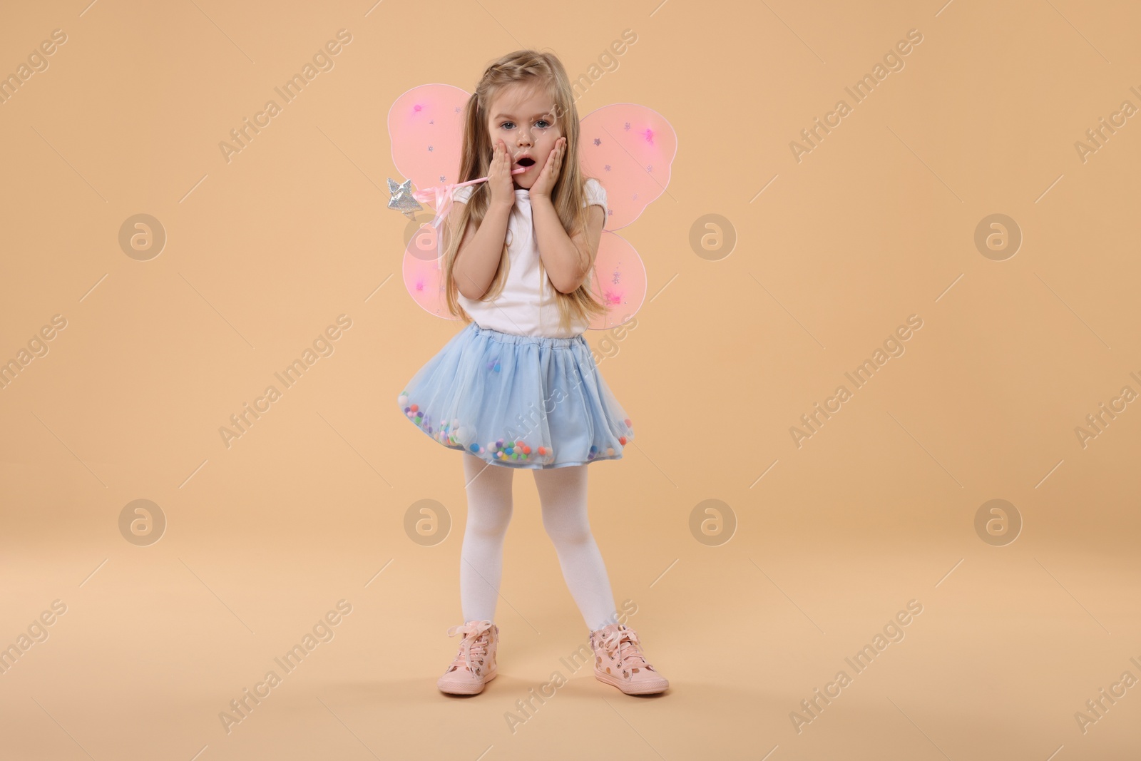 Photo of Surprised little girl in fairy costume with pink wings and magic wand on beige background