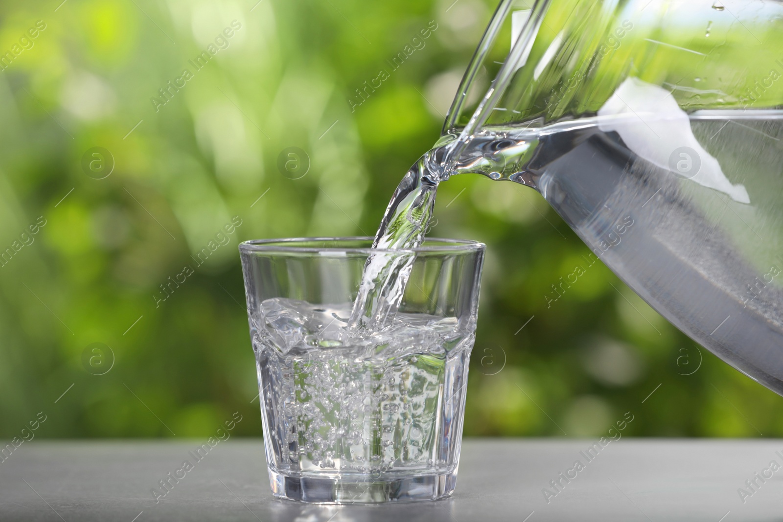 Photo of Pouring water from jug into glass on light grey table outdoors, closeup