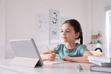 Photo of Little girl doing homework with tablet at table in room