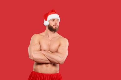 Photo of Attractive young man with muscular body in Santa hat on red background, space for text