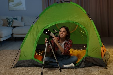 Photo of Young woman using telescope to look at stars while sitting in camping tent indoors