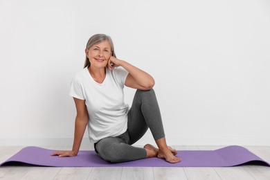 Photo of Happy senior woman sitting on mat near white wall, space for text. Yoga practice