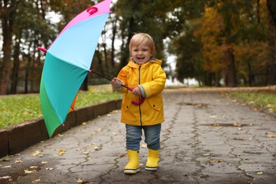 Photo of Little girl holding colorful umbrella and walking near puddles outdoors