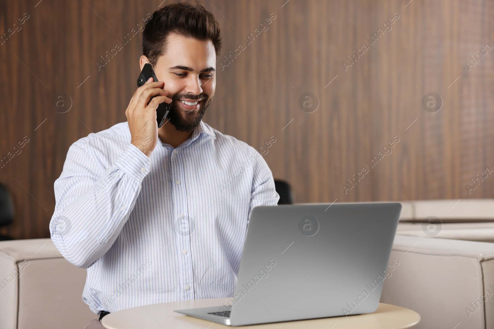 Photo of Happy man using modern laptop while talking on smartphone at table in office