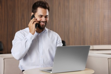 Happy man using modern laptop while talking on smartphone at table in office