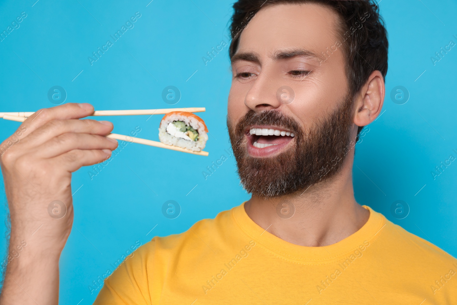 Photo of Happy man eating sushi roll with chopsticks on light blue background