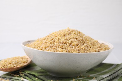 Photo of Bowl and spoon with raw bulgur on table against white background, closeup