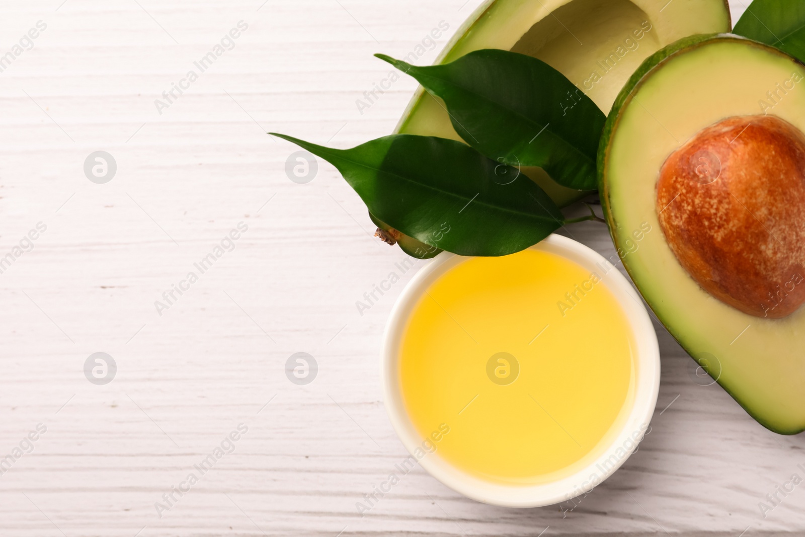 Photo of Cooking oil in bowl and fresh avocados on white wooden table, flat lay. Space for text